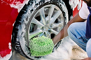 Hands of African man holding green sponge, washing car wheel with foam. Cleaning of modern rims of luxury red car at