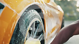 Hands of African man holding yellow sponge, washing car wheel with foam. Cleaning of modern rims of luxury yellow car at