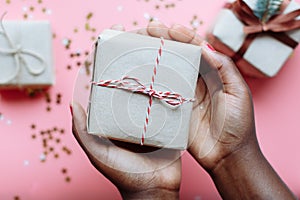 Hands of African woman holding present box with a bow on pastel pink background with stars and snowflakes confetti
