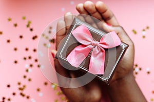 Hands of African woman holding present box with a bow on pastel pink background with stars and snowflakes confetti