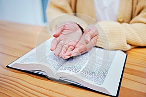 Hands, bible and prayer at desk, religion and Christian worship in home at table. Closeup, holy book and woman in