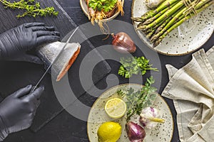 Hands in black gloves cut trout fish on black stone cutting board surrounded herbs, onion, garlic, asparagus, shrimp, prawn in