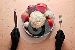 Hands in black gourmet gloves holding fork and knive upon dish full of unprepared vegetables, top view, close-up