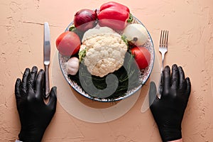 Hands in black gourmet gloves holding fork and knive upon dish full of unprepared vegetables, top view, close-up