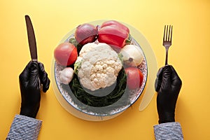 Hands in black gourmet gloves holding fork and knive upon dish full of unprepared vegetables, top view, close-up, bright