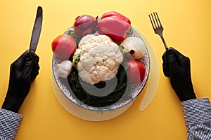 Hands in black gourmet gloves holding fork and knive upon dish full of unprepared vegetables, top view, close-up, bright