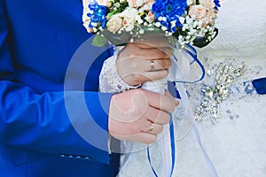 Hands of the bride and groom closeup holding a wedding bouquet of cream roses, blue chrysanthemums and white flowers.