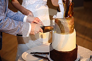 The hands of the bride and groom cut a tiered sweet wedding cake of white gold color. A table with sweets, candy bar.