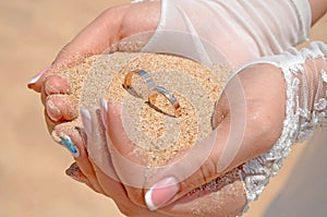 The hands of the bride in white gloves hold a handful of sand and two gold wedding rings.