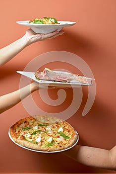 Hands carrying plates with meat tomahawk steak, italian pasta nand cheese pizza. Creative idea, plated in hands