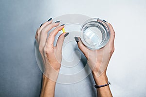 Hands of caucasian girl with yellow pill in left palm and glass of water in right hand.