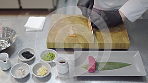 Hands of chef in white restaurant uniform cutting small salmon fish. Kitchenware around, spice, tunafish piece, sauces
