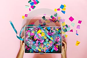 Children`s hands hold a gift box full of colorful confetti. Top view, horizontally on a pink background
