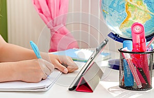 Hands of a child writing homework with tablet and stationery on white desk. Education