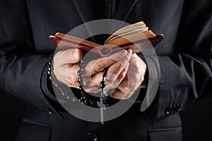 Hands of a christian priest dressed in black holding a crucifix and reading New Testament book