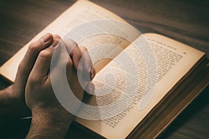 Hands clasped in prayer over a Holy Bible, wooden table background