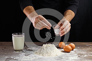 Hands of the cook close-up, breaks the egg into flour, a glass of fresh milk, cooking dough on a dark background
