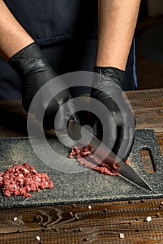 Hands of the cook in gloves cut the red meat on the board. nearby are tomatoes, onion, butter