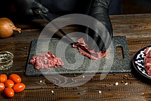 Hands of the cook in gloves cut the red meat on the board. nearby are tomatoes, onion, butter