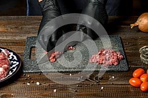 Hands of the cook in gloves cut the red meat on the board. nearby are tomatoes, onion, butter