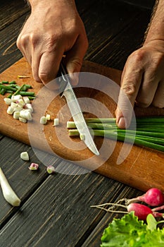 The hands of a cook with a knife cut onions on a cutting board for cooking vegetarian food. Peasant products on the kitchen table