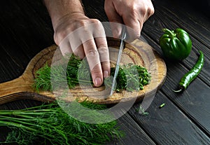 The hands of the cook with a knife are cutting dill on a cutting board for cooking vegetarian food. Peasant products on the