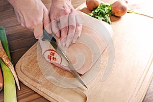 Hands of the cook with a sharp kitchen knife removing the control stamp from the rind of a raw roast pork on a wooden cutting