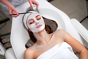 Hands of cosmetology specialist applying facial mask using brush, making skin hydrated and healthy. Attractive woman