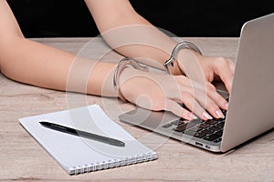 Hands cuffed on the keyboard of a laptop against the background of a Notepad with a pen. The concept of restricting journalistic