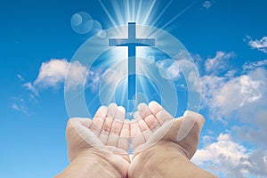 Hands cupped with a shining white cross hovering above on a sunny blue daytime sky