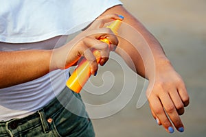 Hands of a dark-skinned woman close up holding a bottle of sunscreen spray applying spf cream on arms and shoulders on a
