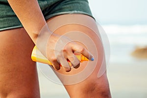 Hands of a dark-skinned woman close up holding a bottle of sunscreen spray applying spf cream on legs and feet on a