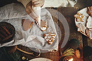 Hands decorating gingerbread cookie christmas tree with icing on rustic table with napkin, spices, candle, decorations. Top view.