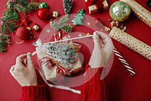 Hands decorating stylish christmas gift with gingerbread cookie and golden paper, ribbon and festive decorations on red background