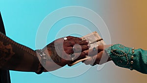 Hands of eastern woman with many rings giving money tied up with twine to seller man, isolated on yellow and blue