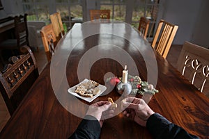 Hands of an elderly man are breaking a cookie at a big family table with candle, Christmas decoration and many empty chairs,