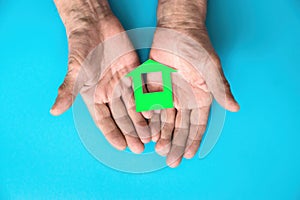 The hands of an elderly man hold a green house cut out of paper on a blue background. Theme of ecology, clean house. The pensioner