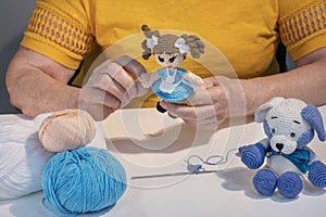 The hands of an elderly woman holding a knitted doll toy. Knitted teddy bear toy and balls of yarn on the table