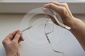 Hands of elderly woman leaning on white windowsill hold thermometer with high temperature readings and glasses in beige frames