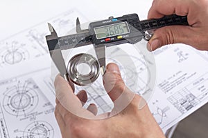 Hands of an engineer measures a metal part with a digital vernier caliper against the background of technical drawings. Quality