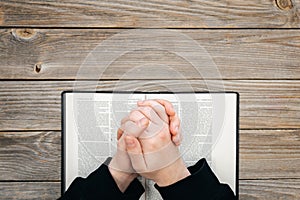 Hands folded in prayer on a Holy Bible on a wooden table, top view.