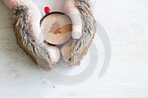 Hands in fur gloves holding mug of hot cocoa on a white wooden background