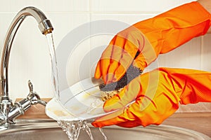 Hands in gloves with sponge wash dirty dishes under running water