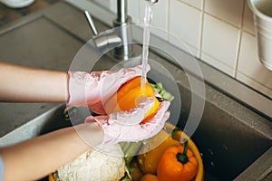 Hands in gloves washing pepper under water stream in sink during virus epidemic. Washing vegetables. Woman in pink hands cleaning