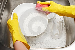 Hands in gloves washing white plate with pink cleaning sponge