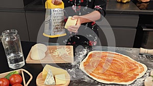 Hands grate cheese on metal grater. Grated cheese in kitchen. Woman making cheese pizza at home. Female preparing homemade food in