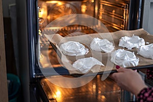 Hands guiding a batch of fresh meringues into the warm glow of an oven, a scene well suited for dessert recipe tutorials