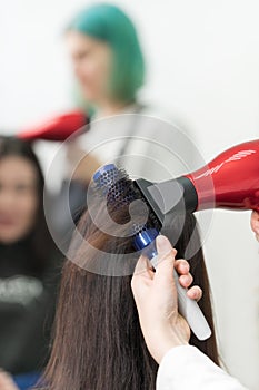 Hands of hairstylist drying brunette hair of customer using red hair dryer and blue comb in beauty salon