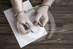 Hands in handcuffs write a turnout on the background of a wooden table.