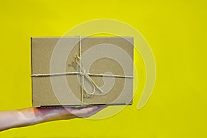Hands hold a cardboard box against a yellow wall. Gift or packaging made of kraft paper, tied criss-cross with jute rope, on a yel photo